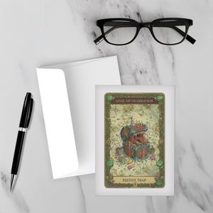 May include: Greeting card with a fantasy illustration of a treasure chest with a monster inside. The card has the text "Level Up Celebration" and "Festive Trap". A black pen, glasses, and an envelope are also in the image.