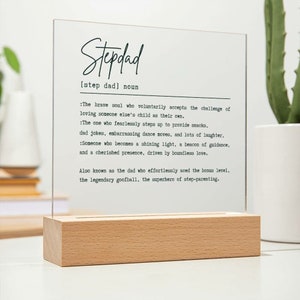May include: A white acrylic sign with a wooden base, featuring a definition of the word "Stepdad". The definition reads: "The brave soul who voluntarily accepts the challenge of loving someone else's child as their own. The one who fearlessly steps up to provide snacks, dad jokes, embarrassing dance moves, and lots of laughter. Someone who becomes a shining light, a beacon of guidance, and a cherished presence, driven by boundless love. Also known as the dad who effortlessly aced the bonus level, the legendary goofball, the superhero of step-parenting."