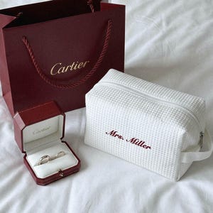 Puede incluir: Una bolsa de papel Cartier color burdeos con letras doradas, un joyero Cartier a juego del mismo color que contiene dos anillos de plata y un neceser rectangular blanco con textura de gofre bordado con el nombre Mrs. Miller en color burdeos.