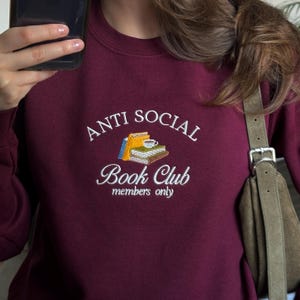Op de afbeelding: Een bordeauxrood sweatshirt met ronde hals en witte geborduurde tekst met de tekst ANTI SOCIAL Book Club members only. Gecentreerd tussen de tekst staat een kleine, kleurrijke geborduurde afbeelding van een stapel van drie boeken met een kopje.