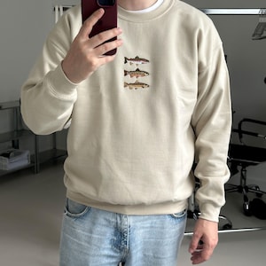 May include: A beige crewneck sweatshirt featuring an embroidered design of three stacked fish on the chest. The sweatshirt has long sleeves with ribbed cuffs and a ribbed hem. The text "MODEL 180 CM (5'11") SIZE L" is printed in white at the bottom left.