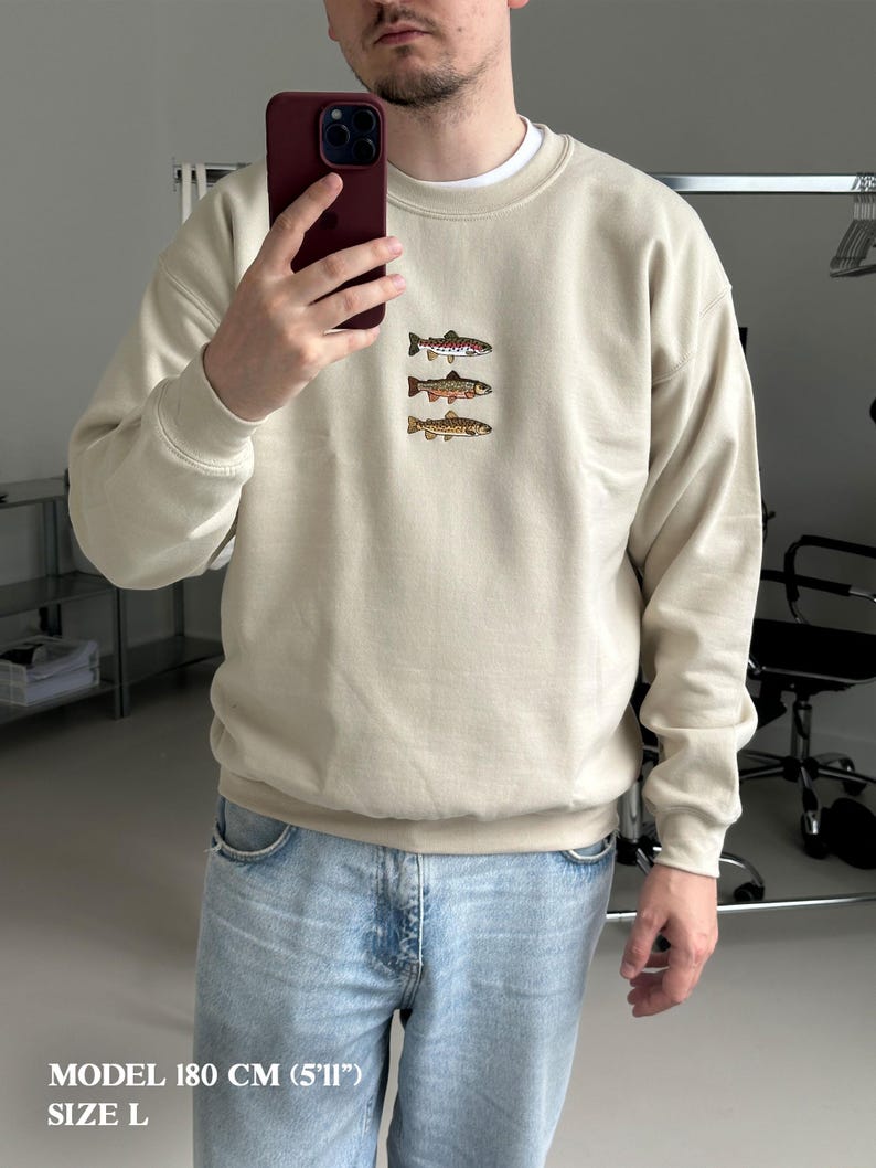 May include: A beige crewneck sweatshirt featuring an embroidered design of three stacked fish on the chest. The sweatshirt has long sleeves with ribbed cuffs and a ribbed hem. The text "MODEL 180 CM (5'11") SIZE L" is printed in white at the bottom left.