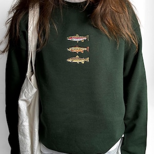 May include: A dark forest green crewneck sweatshirt featuring an embroidered design of three fish stacked vertically on the chest. The fish are depicted in shades of pink, orange, and brown. Text at the bottom reads: MODEL 168 CM (5'6") SIZE M.