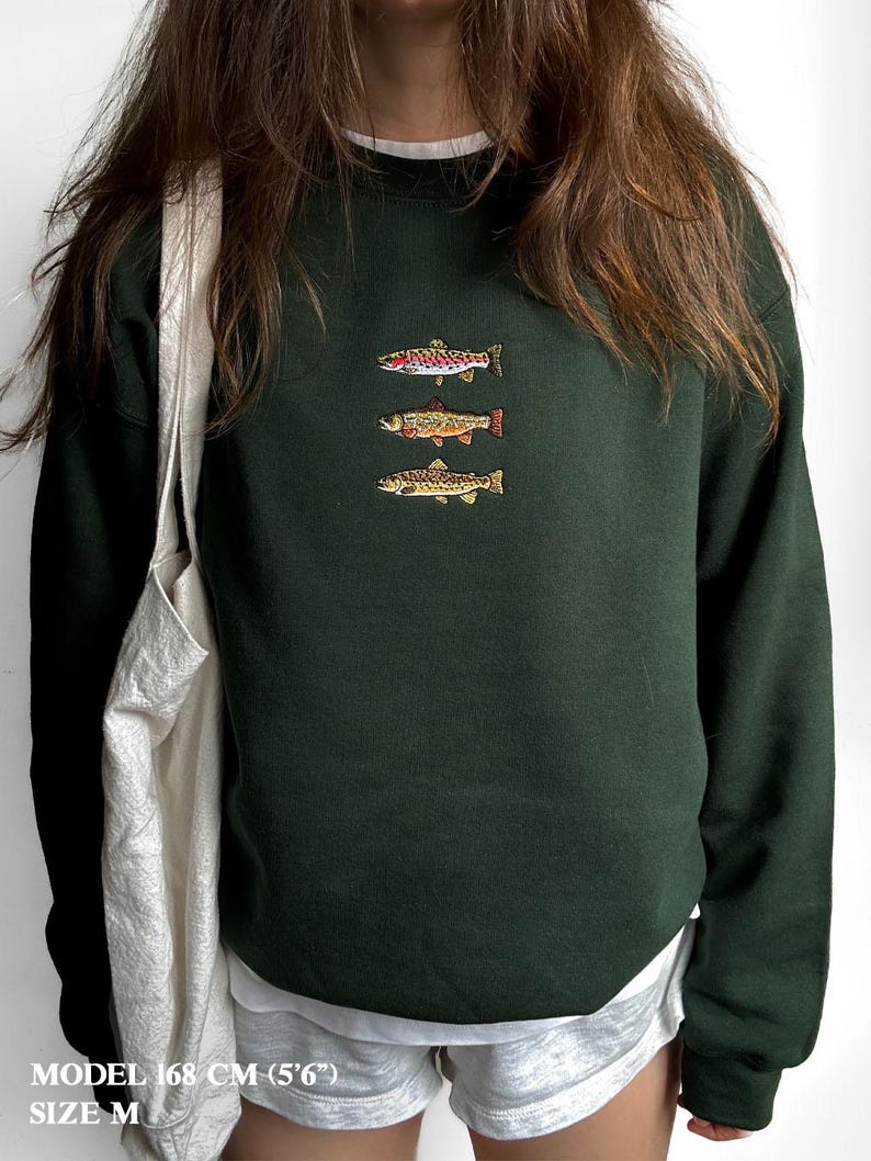 May include: A dark forest green crewneck sweatshirt featuring an embroidered design of three fish stacked vertically on the chest. The fish are depicted in shades of pink, orange, and brown. Text at the bottom reads: MODEL 168 CM (5'6") SIZE M.