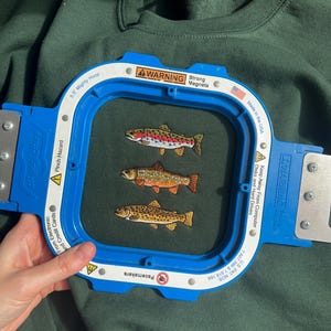 May include: A dark green sweatshirt featuring an embroidery of three fish in a row, secured within a blue 5.5 inch Mighty Hoop embroidery frame. The frame includes white warning labels with text regarding strong magnets and pinch hazards.