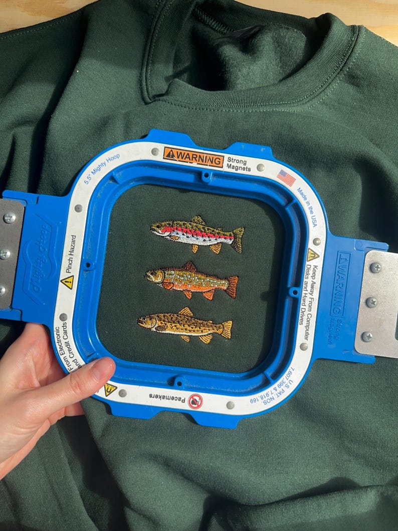 May include: A dark green sweatshirt featuring an embroidery of three fish in a row, secured within a blue 5.5 inch Mighty Hoop embroidery frame. The frame includes white warning labels with text regarding strong magnets and pinch hazards.
