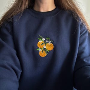 Close-up of a navy embroidered crewneck sweatshirt featuring three orange fruits with green leaves and delicate white blossoms in a botanical floral design.