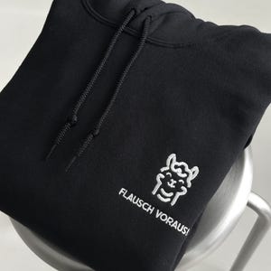 Pode incluir: Uma camisola com capuz preta com um gr&aacute;fico bordado a branco de uma lhama sorridente e o texto "FLAUSCH VORAUS!". A pe&ccedil;a inclui um capuz a condizer com cord&otilde;es pretos e um bolso tipo canguru na parte da frente.