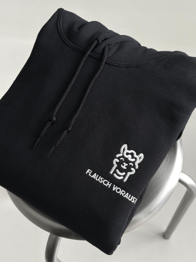 Pode incluir: Uma camisola com capuz preta com um gr&aacute;fico bordado a branco de uma lhama sorridente e o texto "FLAUSCH VORAUS!". A pe&ccedil;a inclui um capuz a condizer com cord&otilde;es pretos e um bolso tipo canguru na parte da frente.