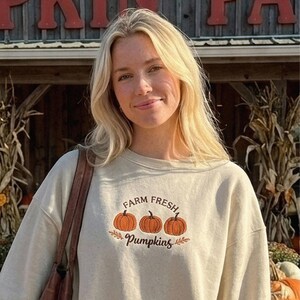 Pode incluir: Uma camisola bege de gola redonda com um design bordado de três abóboras cor de laranja com caules castanhos. Acima das abóboras, o texto "FARM FRESH" está escrito em castanho e, abaixo, a palavra "pumpkins" em letra cursiva castanha.