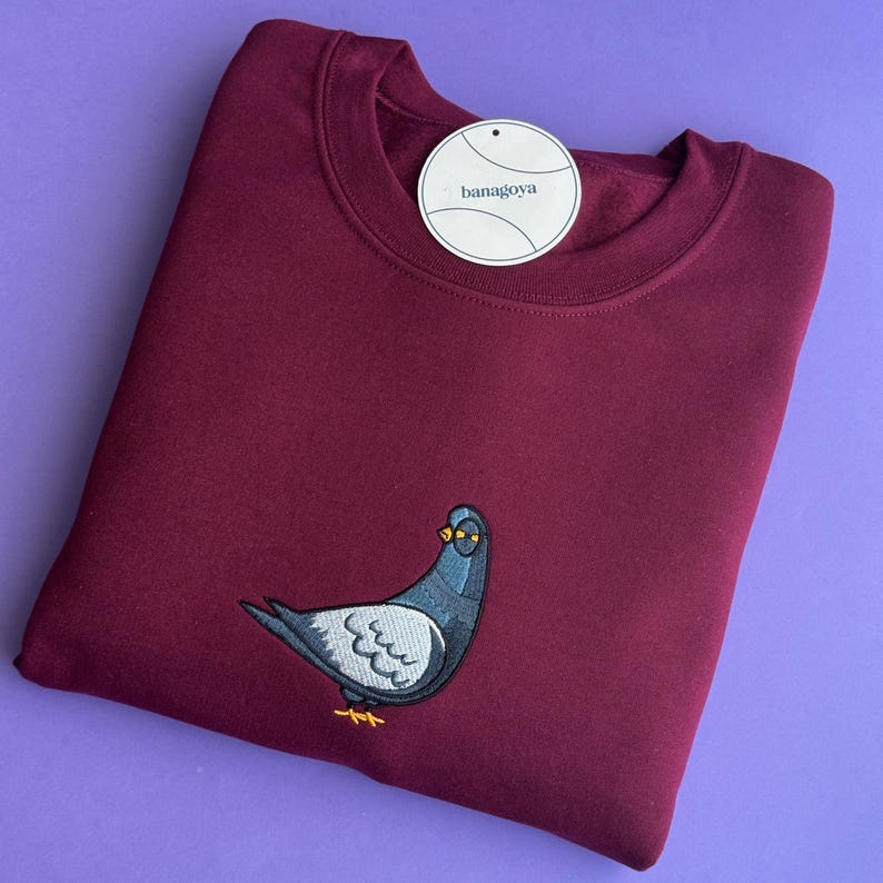 May include: A maroon crewneck sweatshirt featuring an embroidered pigeon wearing yellow glasses on the front. A round white tag with the text banagoya is attached to the neckline. The garment has long sleeves and a ribbed collar.