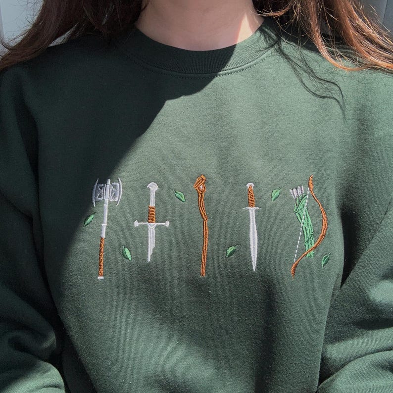 Forest green embroidered Lord of the Rings sweatshirt with mini LOTR weapon embroidery, fantasy inspired crewneck, cozy Middle-earth aesthetic sweater, embroidered Aragorn sword, axe and Legolas bow design.