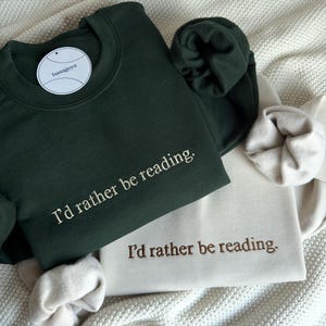Puede incluir: Una sudadera de color verde bosque oscuro y una sudadera de color crema, ambas con el texto bordado "I'd rather be reading." La sudadera verde oscuro incluye una etiqueta de marca blanca circular con el texto "banagoya" cerca del cuello.