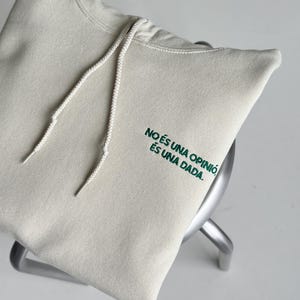 Pode incluir: Uma camisola com capuz de cor creme, com um capuz com cord&atilde;o e texto bordado a verde escuro no peito que diz: NO &Eacute;S UNA OPINI&Oacute;, &Eacute;S UNA DADA.
