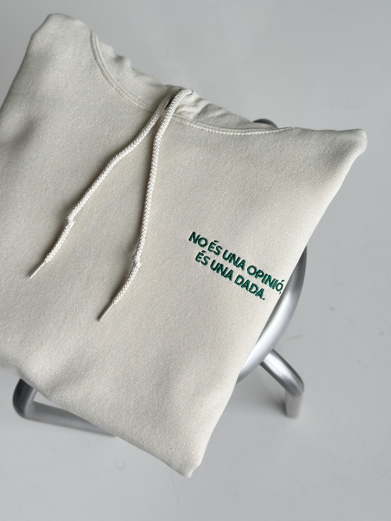 Pode incluir: Uma camisola com capuz de cor creme, com um capuz com cord&atilde;o e texto bordado a verde escuro no peito que diz: NO &Eacute;S UNA OPINI&Oacute;, &Eacute;S UNA DADA.