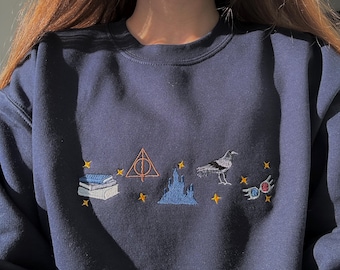 May include: A navy blue crewneck sweatshirt featuring embroidered designs across the chest. The embroidery includes a stack of books, an orange triangle symbol, a blue castle, a black and white bird, and colorful glasses, all accented by small yellow stars.