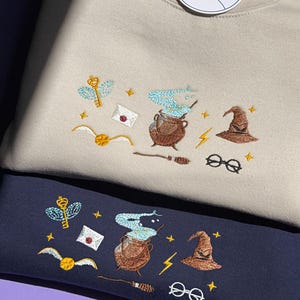 Peut inclure: Un sweat-shirt beige et un sweat-shirt bleu marine, chacun orné de broderies représentant une clé ailée, une lettre, un chaudron avec de la vapeur bleue, un vif d'or, un éclair, un balai, un chapeau et des lunettes rondes. Une étiquette banagoya est fixée.