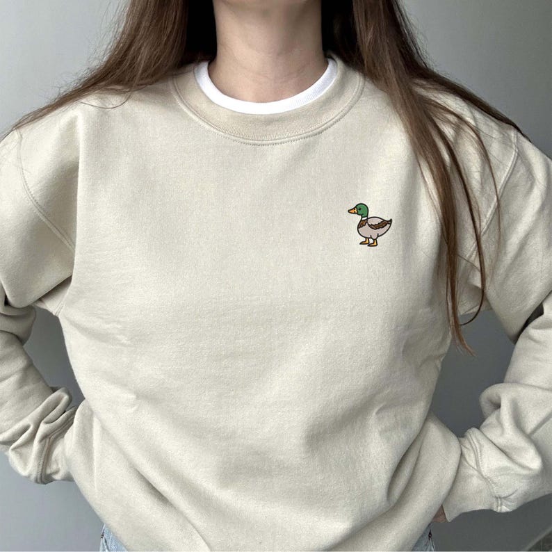 K&ouml;nnte beinhalten: Ein beigefarbenes Sweatshirt mit Rundhalsausschnitt und einer kleinen gestickten Stockente auf der linken Brust. Die Ente hat einen gr&uuml;nen Kopf, einen gelben Schnabel sowie braune und graue K&ouml;rperfedern. Ein wei&szlig;er Kragen ist sichtbar.