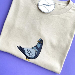 May include: A beige crew neck t-shirt featuring an embroidered pigeon graphic on the front. The pigeon is depicted in shades of blue, grey, and black with yellow feet. A round white tag with the text banagoya is attached to the neckline.