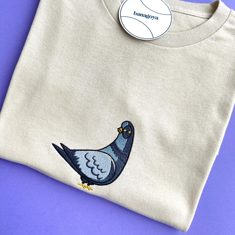 May include: A beige crew neck t-shirt featuring an embroidered pigeon graphic on the front. The pigeon is depicted in shades of blue, grey, and black with yellow feet. A round white tag with the text banagoya is attached to the neckline.