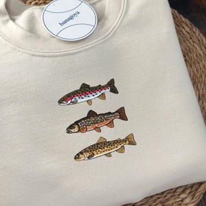 Minimalist embroidered Trout Grand Slam design featuring brook trout, brown trout, and rainbow trout stitched on a cream sweatshirt, perfect fishing shirt for anglers and fly fishing enthusiasts.