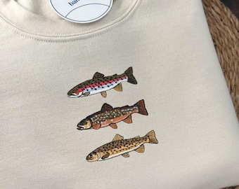 May include: A beige crewneck sweatshirt featuring an embroidered design of three distinct fish stacked vertically. A circular white tag with the text banagoya is attached to the neckline. The fabric has a smooth texture and a ribbed collar.