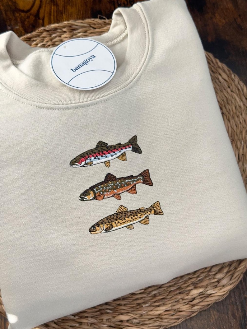 Minimalist embroidered Trout Grand Slam design featuring brook trout, brown trout, and rainbow trout stitched on a cream sweatshirt, perfect fishing shirt for anglers and fly fishing enthusiasts.