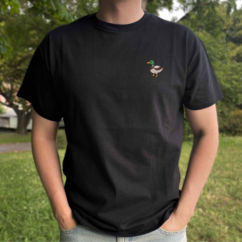 K&ouml;nnte beinhalten: Ein schwarzes T-Shirt mit kurzen &Auml;rmeln und einer kleinen Stickerei einer Stockente auf der linken Brustseite. Die Ente hat einen gr&uuml;nen Kopf, einen gelben Schnabel sowie braune und wei&szlig;e Federn. Das T-Shirt hat einen Rundhalsausschnitt.
