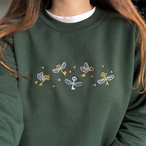 Könnte beinhalten: Ein dunkelgrüner Pullover mit gestickten goldenen Schlüsseln und weißen fliegenden Schlüsseln. Die Schlüssel haben die Form einer Libelle.