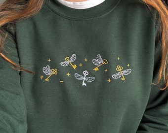 May include: A dark green sweatshirt with embroidered golden keys and white flying keys. The keys are in the shape of a dragonfly.