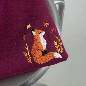 Könnte beinhalten: Ein tiefburgunderfarbenes Sweatshirt mit einer Stickerei eines orangefarbenen Fuchses mit weißer Brust und weißer Schwanzspitze. Der Fuchs ist von gestickten gelben und orangefarbenen Herbstblättern sowie kleinen Zweigen umgeben.