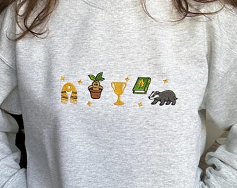 May include: A light gray crewneck sweatshirt featuring embroidered icons across the chest, including a yellow and black striped scarf, a potted mandrake, a gold trophy, a green book with a leaf emblem, and a badger, accented by small yellow stars.