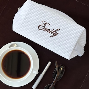 Könnte beinhalten: Eine weiße, strukturierte Kosmetiktasche mit dem Namen "Emily" in Braun gestickt. Die rechteckige Tasche hat einen Reißverschluss. Eine Tasse Kaffee, Mascara und Sonnenbrille liegen auf einer braunen Oberfläche.