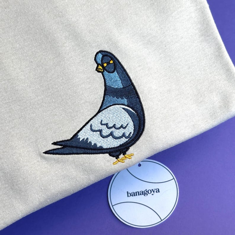 May include: A light gray t-shirt featuring an embroidered pigeon graphic in shades of blue, dark gray, and yellow. The pigeon is depicted in profile with a yellow beak and feet. A round white tag with the black text banagoya is attached.