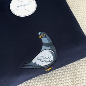 May include: A dark navy blue sweatshirt featuring an embroidered pigeon graphic in shades of grey, blue, and yellow. A round white tag with the text banagoya is attached to the upper left corner of the garment, which has a smooth fabric texture.
