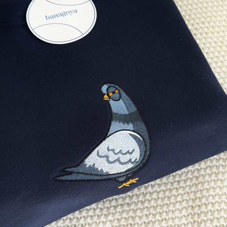 May include: A dark navy blue sweatshirt featuring an embroidered pigeon graphic in shades of grey, blue, and yellow. A round white tag with the text banagoya is attached to the upper left corner of the garment, which has a smooth fabric texture.