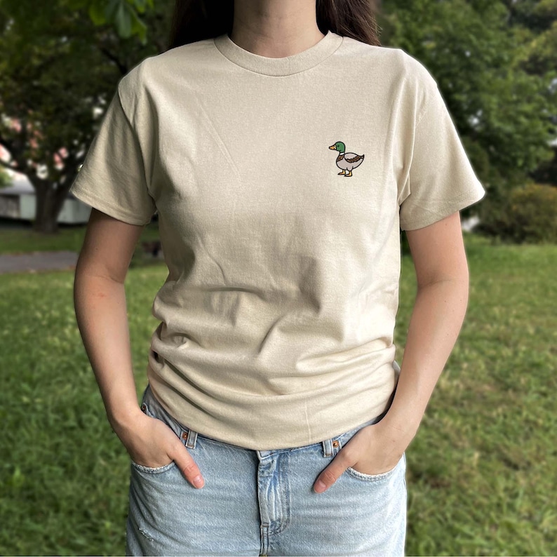 K&ouml;nnte beinhalten: Ein beiges Kurzarm-T-Shirt mit Rundhalsausschnitt und einer kleinen gestickten Stockente auf der linken Brust. Die Ente hat einen gr&uuml;nen Kopf, einen gelben Schnabel sowie braune und wei&szlig;e Federn. Das T-Shirt wird mit einer hellblauen Jeans kombiniert.