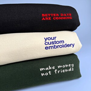 Three folded fabric swatches with embroidered text. The top swatch is black with red text that reads "Better Days Are Coming". The middle swatch is cream with blue text that reads "your custom embroidery". The bottom swatch is dark green with white text that reads "make money not friends".