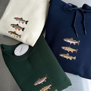 May include: Three long-sleeve garments in cream, dark green, and navy blue, each featuring an embroidered design of three stacked fish. The navy blue garment includes a hood with matching drawstrings, while the others are crewneck style.