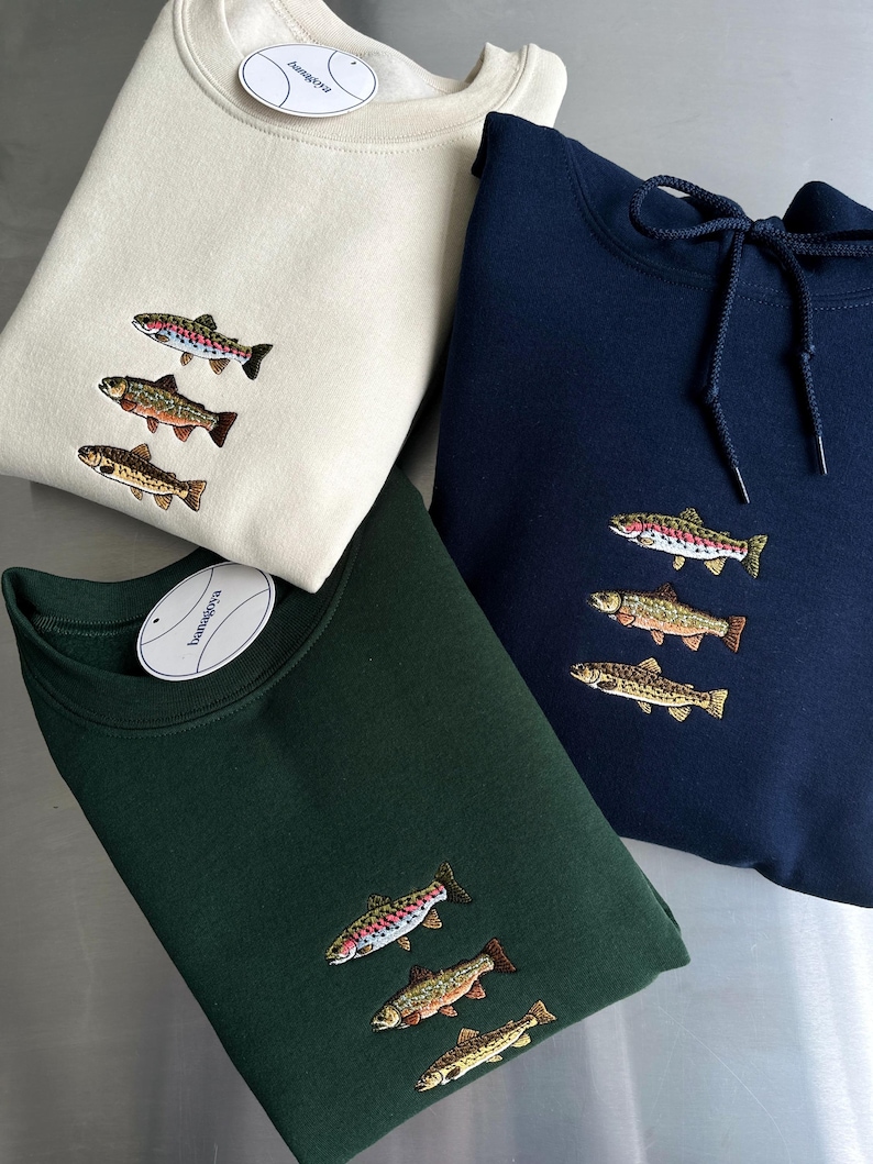 May include: Three long-sleeve garments in cream, dark green, and navy blue, each featuring an embroidered design of three stacked fish. The navy blue garment includes a hood with matching drawstrings, while the others are crewneck style.