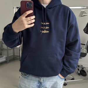 May include: A navy blue hooded sweatshirt featuring an embroidered graphic of three stacked fish on the chest. The hoodie includes a front kangaroo pocket and matching drawstring cords. The model is 180 cm (5'11") tall and wears a size XL.