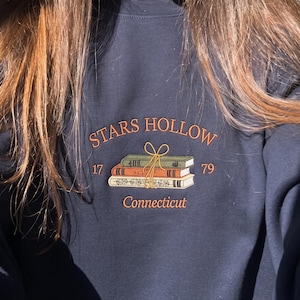 Könnte beinhalten: Ein marineblaues Sweatshirt mit Rundhalsausschnitt und einer Stickerei von drei gestapelten Büchern, die mit einem gelben Band zusammengebunden sind. Der Text "STARS HOLLOW" steht darüber, "17" und "79" an den Seiten und "Connecticut" in Orange darunter.