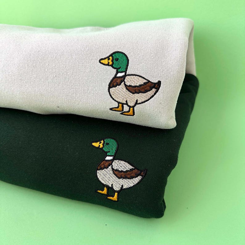 K&ouml;nnte beinhalten: Ein hellbeiges Sweatshirt und ein dunkelwaldgr&uuml;nes Sweatshirt, die &uuml;bereinander gestapelt sind. Jedes Kleidungsst&uuml;ck zeigt ein gesticktes Stockenten-Design mit gr&uuml;nem Kopf, gelbem Schnabel sowie braunen und wei&szlig;en K&ouml;rperdetails auf der Brust.