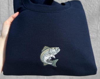 May include: A navy blue crewneck sweatshirt featuring a detailed embroidered graphic of a striped bass fish on the center chest. The fish is depicted in shades of silver, white, and olive green, showcasing its scales and fins in a realistic style.