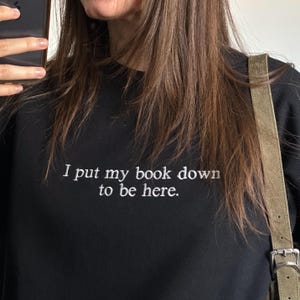 Może przedstawiać: Czarna bluza z długim rękawem z białym haftowanym napisem: I put my book down to be here. Odzież jest noszona z zamszową torbą na ramię w kolorze opalenizny ze srebrną metalową klamrą i regulowanym paskiem.