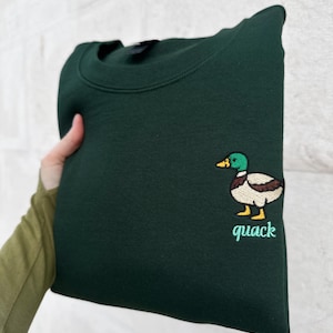 K&ouml;nnte beinhalten: Ein dunkelwaldgr&uuml;nes Sweatshirt mit Rundhalsausschnitt und einer gestickten Stockente auf der Brust. Die Ente hat einen gr&uuml;nen Kopf, eine braune Brust, einen wei&szlig;en K&ouml;rper und gelbe F&uuml;&szlig;e. Unter der Ente ist das Wort quack hellgr&uuml;n gestickt.
