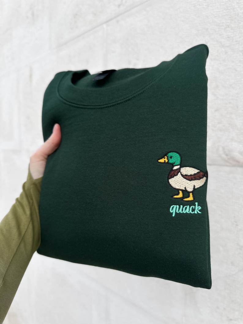 K&ouml;nnte beinhalten: Ein dunkelwaldgr&uuml;nes Sweatshirt mit Rundhalsausschnitt und einer gestickten Stockente auf der Brust. Die Ente hat einen gr&uuml;nen Kopf, eine braune Brust, einen wei&szlig;en K&ouml;rper und gelbe F&uuml;&szlig;e. Unter der Ente ist das Wort quack hellgr&uuml;n gestickt.