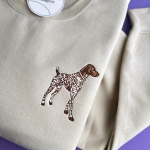 Könnte beinhalten: Beiges Sweatshirt mit einem gestickten braun-weißen Hundemotiv. Der Hund trägt ein Halsband. Ein weißes Etikett mit dem Text "banagoya" ist am Sweatshirt befestigt.