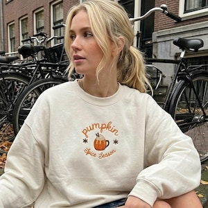 Puede incluir: Una sudadera de cuello redondo color crema con un bordado que muestra una taza con forma de calabaza, nata montada y dos agitadores. El texto "pumpkin spice season" está bordado en letras cursivas de color naranja alrededor del diseño.