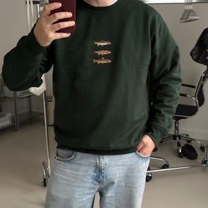 May include: A dark forest green crewneck sweatshirt featuring a small, centered embroidery of three stacked fish in shades of brown, orange, and white. The garment has long sleeves with ribbed cuffs and a ribbed neckline, paired with light blue denim jeans.